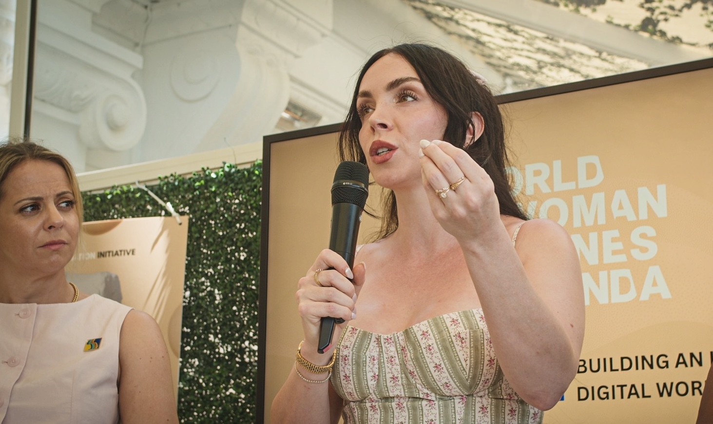 woman with brown hair speaking into microphone and gesturing with left hand